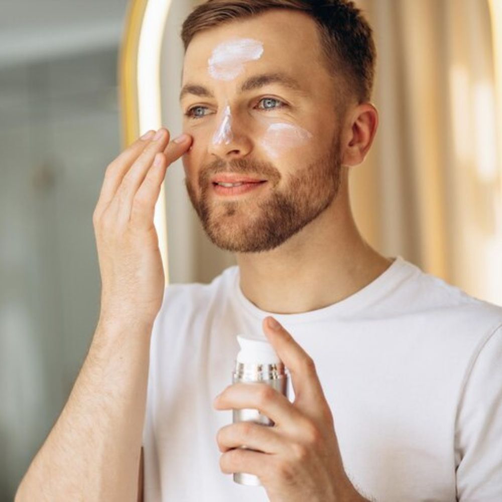 Bearded man applying white face moisturizer cream in bathroom mirror